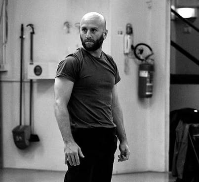 Uomo con capelli rasati e barba, in abbigliamento sportivo, si allena in uno spazio interno.