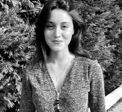 Giovane donna sorridente in primo piano, con capelli lunghi e abbigliamento elegante, sullo sfondo di piante verdi.