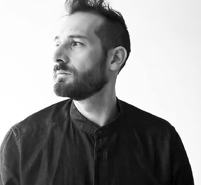 Uomo con barba e capelli corti, indossa una camicia scura e guarda verso sinistra, su sfondo chiaro.