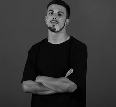 Giovane uomo con braccia incrociate, sguardo serio, indossa una maglietta nera su sfondo grigio.