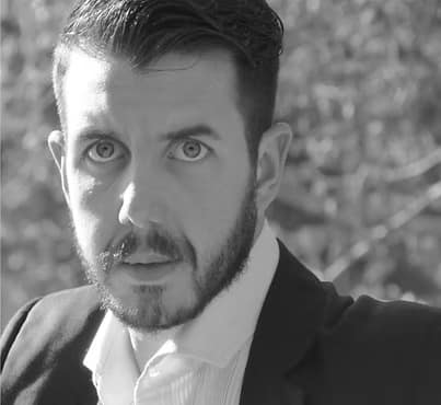 Uomo con barba e capelli scuri, sguardo intenso, indossa un abito elegante su sfondo sfocato.