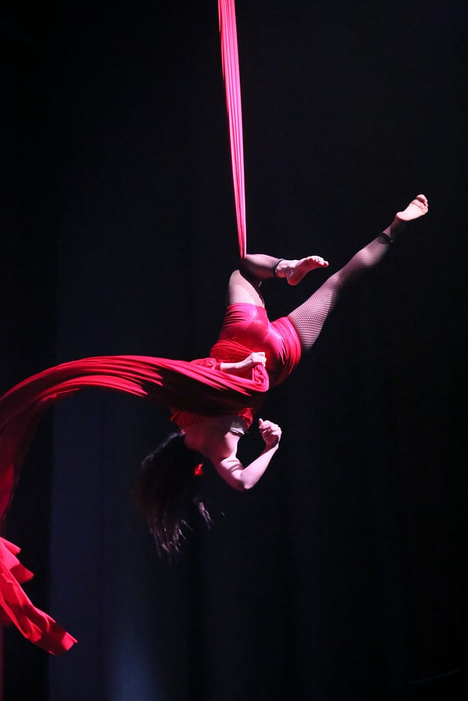 Artista acrobatica sospesa in aria, avvolta in tessuti rossi, mentre esegue una figura elegante.