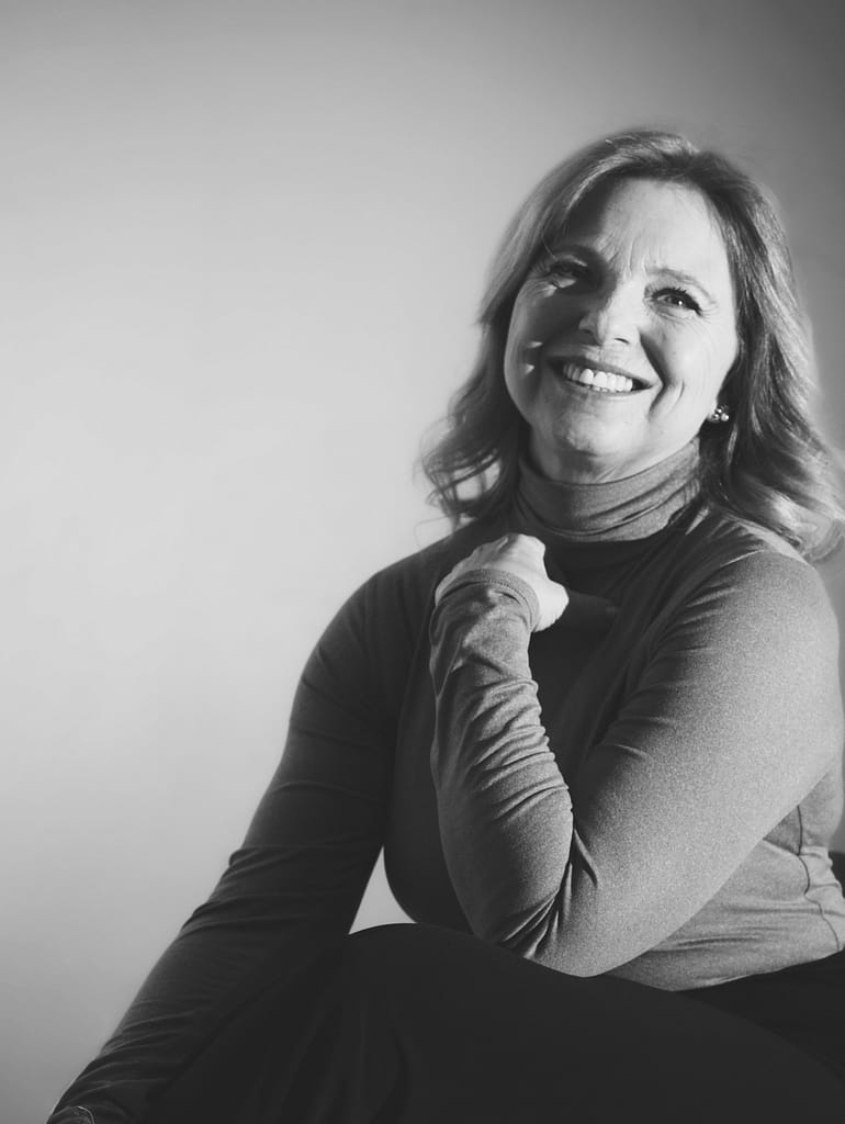 Donna sorridente con capelli lunghi e mossi, indossa un maglione a maniche lunghe e posa con una mano sul collo.