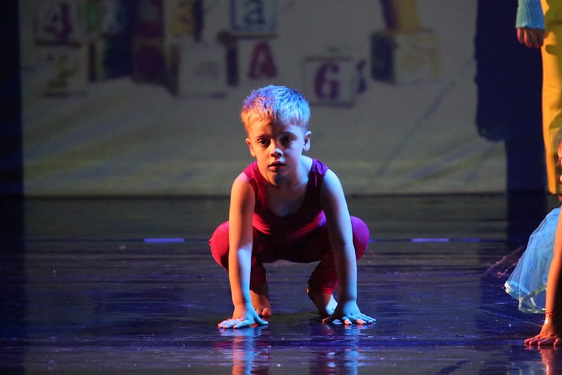 Bambino in posizione accovacciata su un palco, con abbigliamento rosso e capelli biondi, in un contesto di danza.