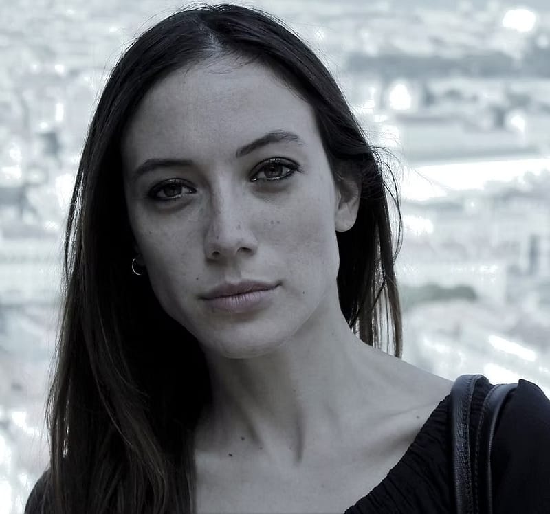 Donna con capelli lunghi e scuri, sguardo intenso, sfondo urbano sfocato.